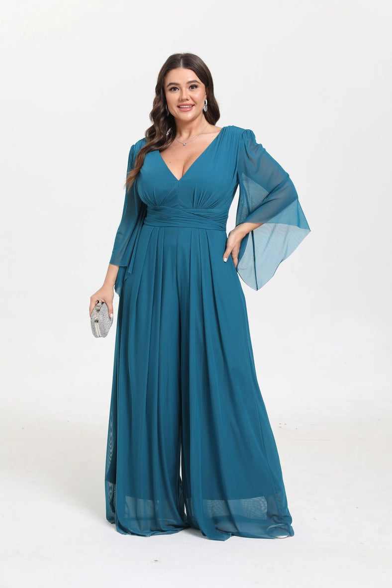 A woman modeling a floor-length teal blue maxi dress with a V-neckline, sheer three-quarter sleeves with wide flared cuffs, and a flowing skirt, holding a small silver clutch against a plain white studio background.